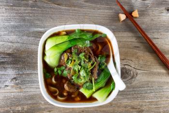 High angled view of beef noodle soup with bok choy and parsley. Soup in soup with chopsticks in holder on rustic wood.  