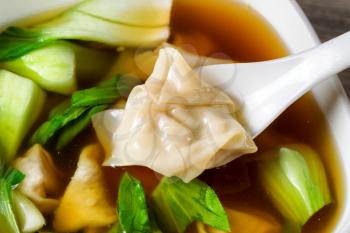 Close up Chinese wanton soup, single piece in spoon, with bok choy on rustic wood. 