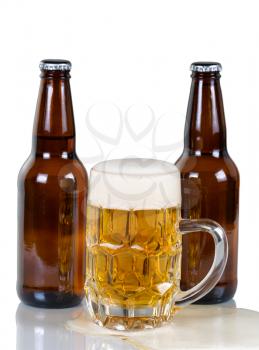 Golden colored beer flowing out of glass mug with two full bottles in background. Isolated on white with reflection. 