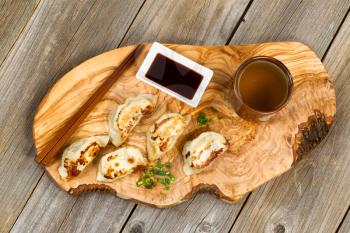 Top view of freshly cooked Chinese dumplings, green tea, soy sauce, sprinkle of green onions and chopsticks resting on olive wooden server board. 