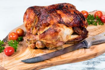 Close up front view of barbecued chicken with vegetables, herbs and large cutting knife on wooden server with white marble underneath. Selective focus on upper front leg of chicken. 
