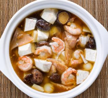 High angled view of Asian soup consisting of shrimp, mushroom, tofu, and vegetable on bamboo mat. 