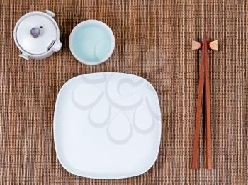 Overhead view of chopsticks, plate, cup and tea server on bamboo mat.  