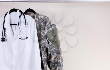 Medical Doctor Consultation white coat, stethoscope around collar, and military uniform hanging on rack. Off white wall in background. Horizontal layout with copy space. 
