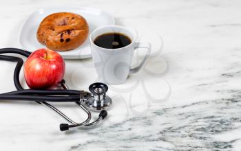 Medical stethoscope with apple, dark coffee, and bagel on white marble. Selective focus on stethoscope. Healthy breakfast concept. 