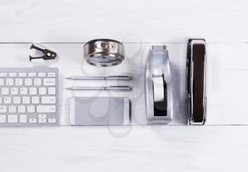 Overhead view of stainless steel stationery items consisting of stapler, tape dispenser, pens, stapler remover, cell phone, keyboard and clock on white wood. 