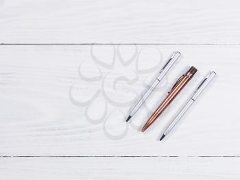 Overhead view of three metal pens on white wood. 