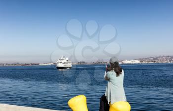 Back view of a woman taking photos with cell phone of San Diego bay in Southern California while sitting down at pier. 