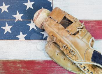 Old worn baseball mitt and baseball on faded wooden boards painted red, white and blue with flag pattern. 