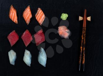Overhead view of fresh Japanese sushi, ginger, wasabi, and chopsticks on black slate. 
