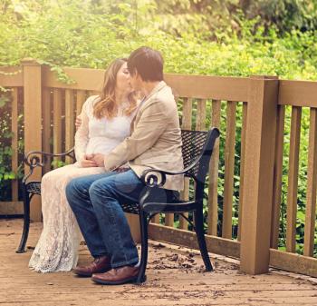 Expecting mom and dad kissing on bench patio with woods in background. Haze light effect applied to image.    