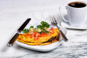 Close up of freshly made omelet with sliced tomato, onion, parsley, and mushroom in dish with silverware on white marble counter. Dark coffee in background.