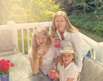 Top front view of happy mother and daughters, looking forward, drinking grapefruit juice while outdoors on patio with woods in background. Light haze effect applied. 