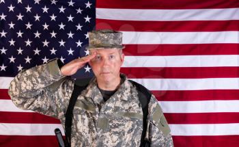 Veteran male soldier, facing forward, saluting with USA flag in background. Soldier armed with military weapon and ammo clips. 
