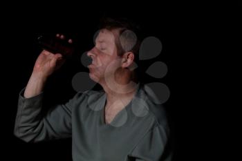 Depressed man drinking beer out of a bottle while sitting down with eyes close in a dark background. 