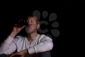 Depressed man drinking beer out of a bottle while sitting down in a dark background. 