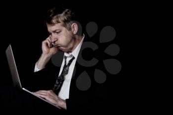 Side view of stressed business man on cell phone while looking at computer screen.  Dark background with light on subject.  