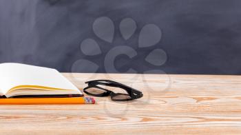 Back to school concept with reading glasses, pencils and notepad on desktop with erased black chalkboard in background. 