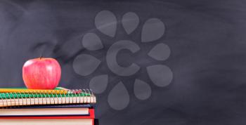 Back to school concept with apple, pencils, spiral notepad and books in front of black chalkboard. 
