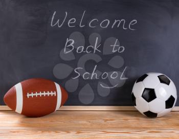 Football and soccer ball on desktop. Erased black chalkboard in background along with welcome back to school message to students. 