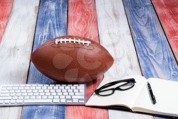 Front view of computer keyboard, reading glasses, notepad, pen and American football on red, white and blue rustic wooden boards. Concept of draft and plays for the season. 