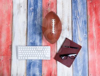 Overhead view of computer keyboard, reading glasses, notepad, pen and American football on red, white and blue rustic wooden boards. Concept of draft day and future plays for the season. 