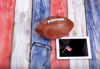 Overhead view of mobile computer, reading glasses, and American football on red, white and blue rustic wooden boards. Concept of using technology for football. 