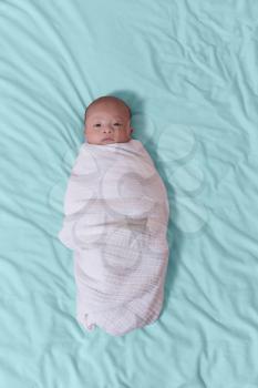 Overhead view of infant baby boy swaddled in white blanket with eyes open while lying on blue sheets. Vertical layout