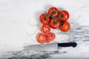 Overhead view of freshly sliced garden tomatoes with large kitchen knife and whole tomatoes on natural marble stone counter. 
