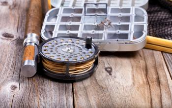 Close up view of antique fly reel, rod, and landing net with lure container on rustic wooden boards. Selective focus on reel. 