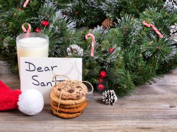Stack of cookies, a glass of milk and letter for Santa with evergreen decoration in background.