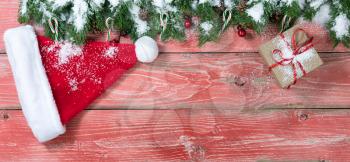Snowy rustic red wooden background for Christmas concept with fir branches, candy canes, red berries, Santa cap, gift box and pine cones. Overhead view with copy space.