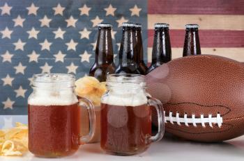 Pint glasses of beer, football, bowl of potato chips, cold beer in bottles on white glass table with United States flag painted on rustic wood. Layout in horizontal format. 