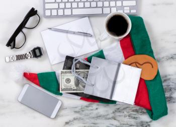 Overhead view of office desktop with computer keyboard, cell phone, shopping bag, wallet, money, pen, paper, reading glasses, coffee and Xmas Stocking on marble surface. Shopping for Christmas concept