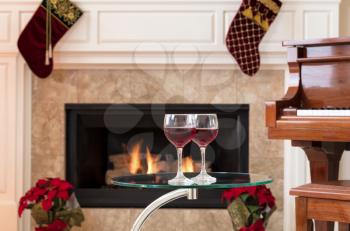 Two glasses of red wine on top of glass table with glowing fireplace, piano and holiday decorations in background 