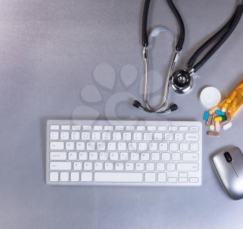 Overhead view of a medical stethoscope, computer keyboard, mouse and pills on stainless steel table