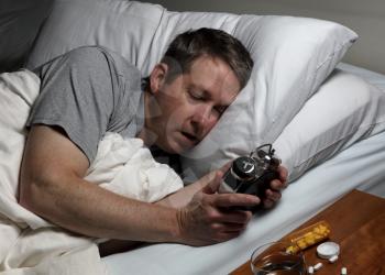 Mature man holding and staring at alarm clock while trying to fall asleep. Insomnia concept. 