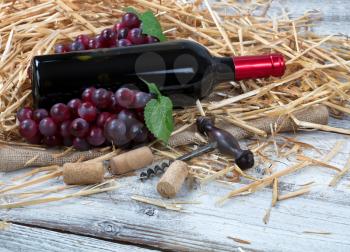 Close up view of a red wine bottle, grapes plus corkscrew with straw and burlap on white rustic boards 