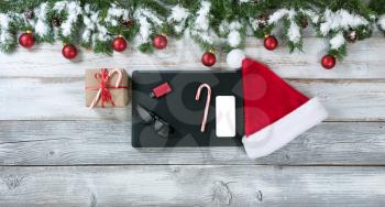 Christmas background with Santa Claus cap, mobile computer equipment and gift with traditional decorations on rustic white wood. 