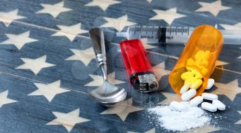 Front view of opioid pain killer tablets with spoon, lighter and syringe on rustic USA flag in background for drug addiction concept in America  