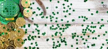 St Patrick day good luck hat, clovers and horseshoe with shiny gold coins forming left border on rustic white wooden boards 