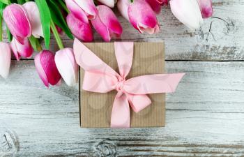 Close up overhead view of a brown gift box with pink tulips in background on white weathered wooden boards 