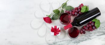 Overhead view of Red wine bottle with grapes, drinking glasses, corkscrew, gift box and rose on natural marble stone