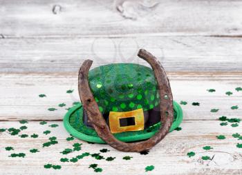 Close up view of a rusty horseshoe resting on hat for St Patrick day with shiny clovers on weathered white wooden boards 