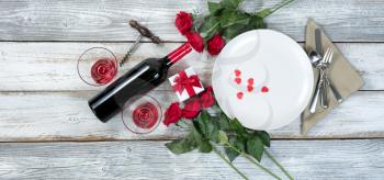 Valentine dinner setting with gift, red roses, wine and heart shapes on rustic table
