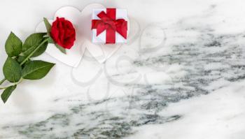 Single red rose resting on card and gift box on marble stone background in flat lay view 