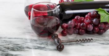 Close up view of a corkscrew with red wine bottle, filled glasses and grapes on natural marble stone setting in background