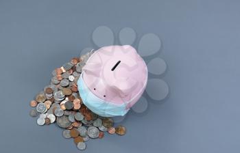 Piggy bank with surgical mask on face with coins on gray desk