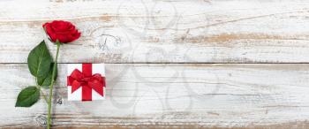 Anniversary background with a single red rose and wrapped gift box on white rustic wood 
