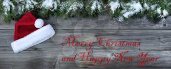 Merry Christmas and New Year concept on rustic natural wooden background with snow covered Fir branches plus Santa Claus cap  and text 